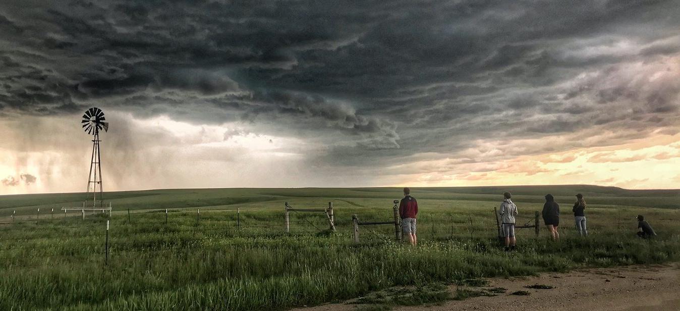 Students in a field with a windmill observing storms over the plains